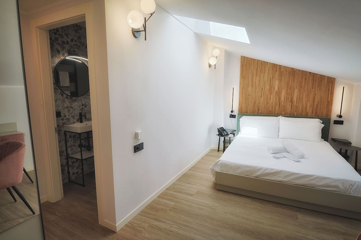 A modern hotel room with a large bed, a wooden headboard, a small desk with a chair, a mirror, and a bathroom with a sink and vanity.