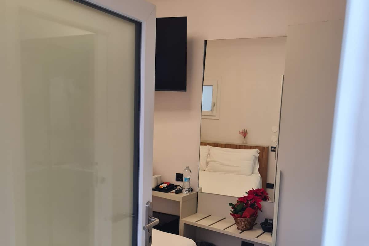 A compact hotel room with a bed, bedside table, and a small desk with a chair. A mirror and a wall-mounted TV are visible.