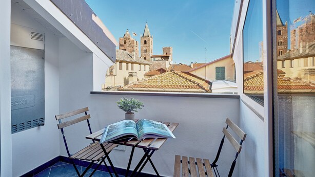 A balcony with a view of historic buildings, a table with an open book, and a potted plant. - Tra le Torri (Albenga)
