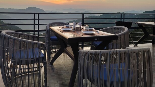 An outdoor dining area with wicker furniture and a table set for two, overlooking a scenic view of mountains and a sunset. - Aagaman- Your Mountain Home (Naldehra)