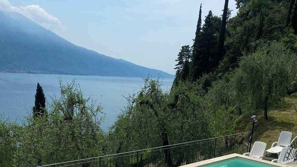 Et rektangulært pool med krystalklart blåt vand, omgivet af en flisbelagt terrasse. Der er hvide liggestole og et lille bord med stole på terrassen. Poolen ligger foran et baggrund af træer og fjerne bjergkæder, under en klar himmel.