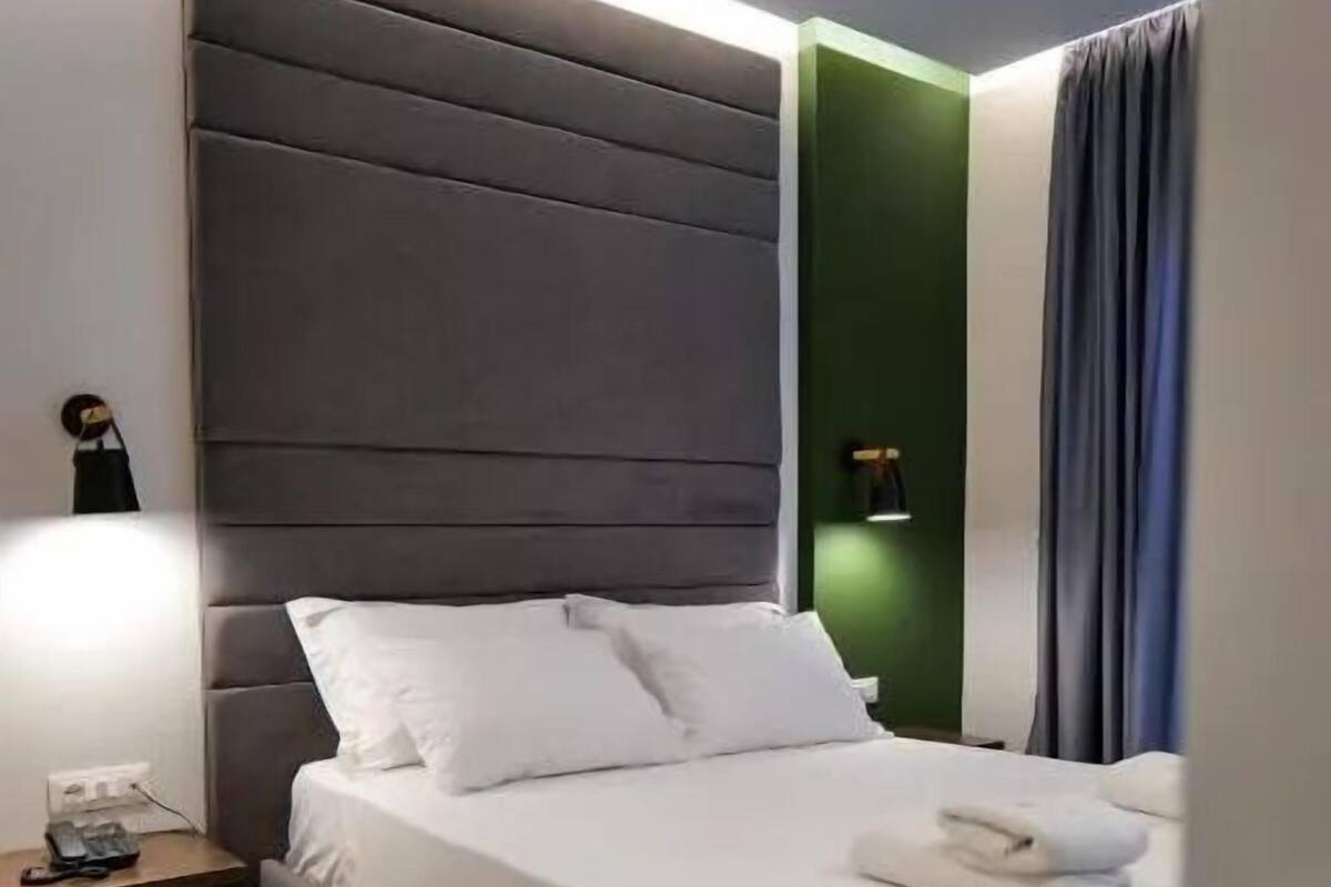 Una cama bien hecha con sábanas blancas, un cabecero tapizado en gris y una mesita de noche con una lámpara.