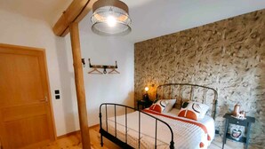 A bedroom with a wooden door, a bed with a metal frame, a stone wall, a hanging light fixture, and a small table with a lamp. - Guest rooms : La Cavalière - LA SOURCE DE NOS RACINES - CORPS DE FERME - AVESNOIS (Beaufort)