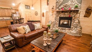 A cozy living room with a stone fireplace, wooden walls, and a leather sofa. - As You Wish Cabin in a breathtaking one-bedroom honeymoon cabin that is the perfect place to make lasting memories with each other. (Broke Bow)