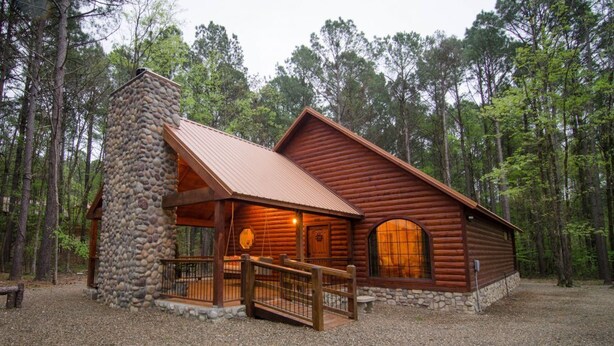 A log cabin with a stone chimney, wooden deck, and large windows. - As You Wish Cabin in a breathtaking one-bedroom honeymoon cabin that is the perfect place to make lasting memories with each other. (Broke Bow)