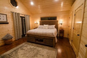 Un dormitorio de cabaña de madera con cama, mesitas de noche, una ventana y una puerta.