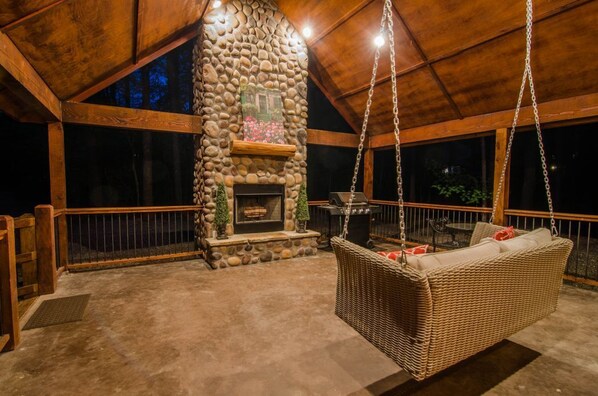 An outdoor patio with a stone fireplace, a hanging wicker swing, and a wooden deck.