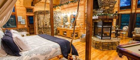 Interior acolhedor de uma cabana de madeira com cama, lareira de pedra e mesa de bilhar.