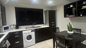 Una cocina moderna con lavadora integrada, una zona de comedor con mesa y sillas, y un ventanal de marco negro con persianas.