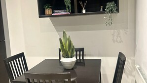 Un comedor con una mesa de madera oscura y cuatro sillas, una planta en maceta sobre la mesa, y un estante negro arriba con libros y objetos decorativos.