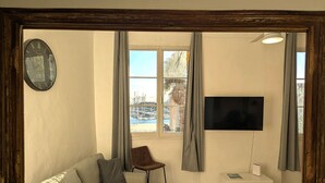 Habitació amb un mirall de fusta que reflecteix una vista de port esportiu, un sofà, una cadira, una televisió de pantalla plana i un petit armari blanc.