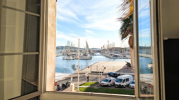 Vista d'una finestra sobre un port esportiu amb vaixells amarrats, una passarel·la per a vianants i vehicles estacionats.