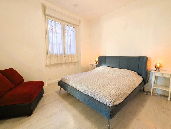 A bedroom with a bed, a nightstand, a red chair, and a window with curtains.