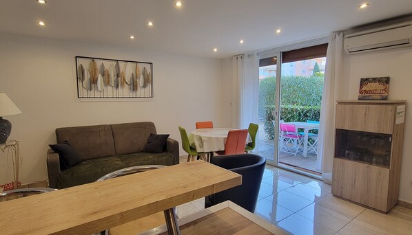 A living room with a sofa, a dining table, and a glass door leading to an outdoor area with a table and chairs.