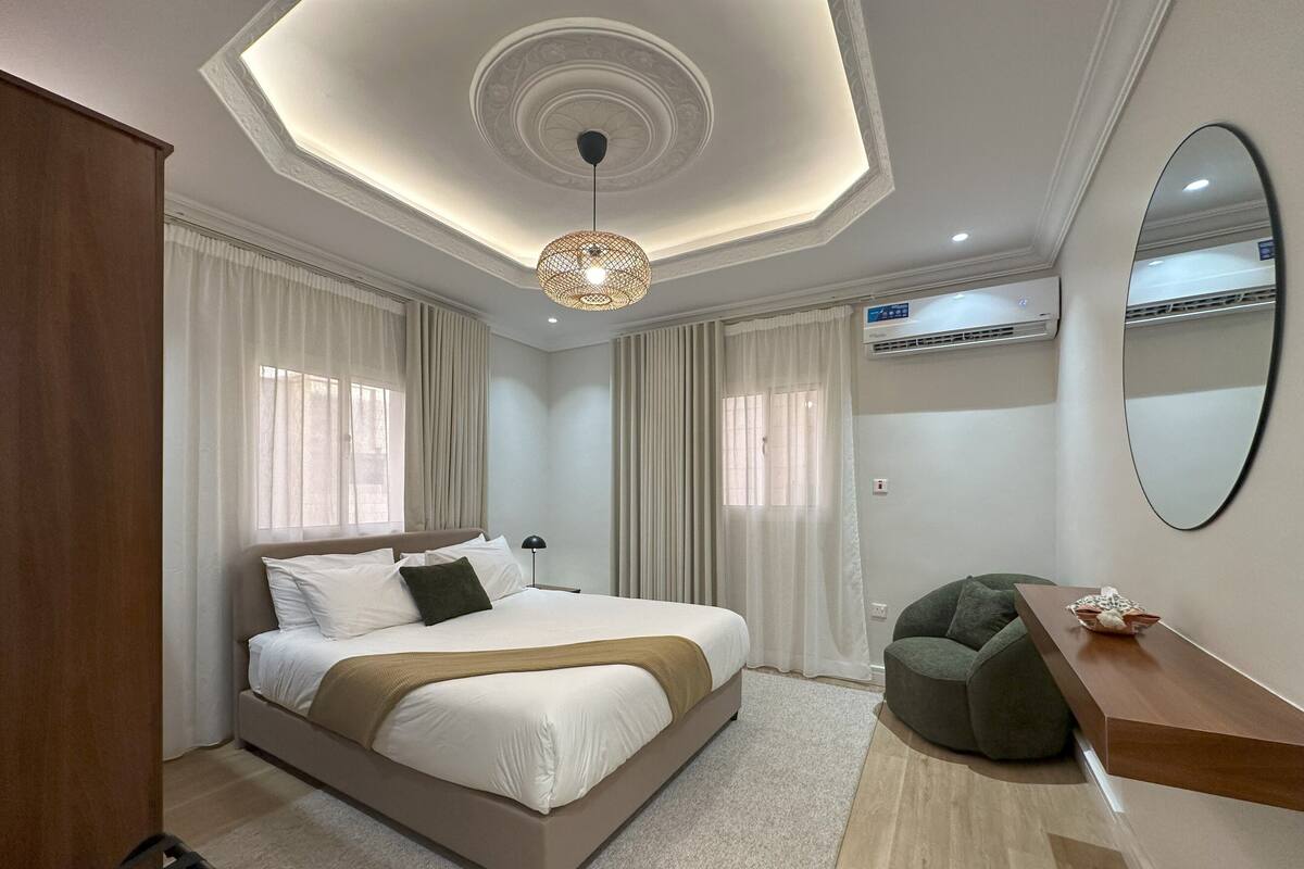 A modern bedroom with a large bed, a wooden wardrobe, a green armchair, a wooden bedside table, and a ceiling light fixture.