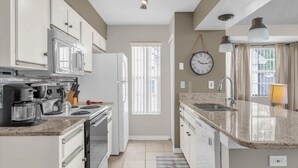 Eine moderne Küche mit weißen Schränken, einem Herd und einer Kaffeemaschine. Ein Fenster mit Jalousien, eine Wanduhr und ein Spülbecken mit Wasserhahn sind ebenfalls vorhanden.