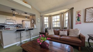Ein Wohnzimmer mit braunem Ledersofa, einem Couchtisch aus Holz mit einer Vase voller rosa Blumen, einer Küchenzeile mit weißen Schränken, einer Mikrowelle und einem Spülbecken.