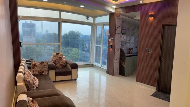 A spacious living area with a sofa set, a kitchen area, and large windows offering a view of the outdoors. - BEST HOME STAY IN VRINDAVAN (Vrindavan)