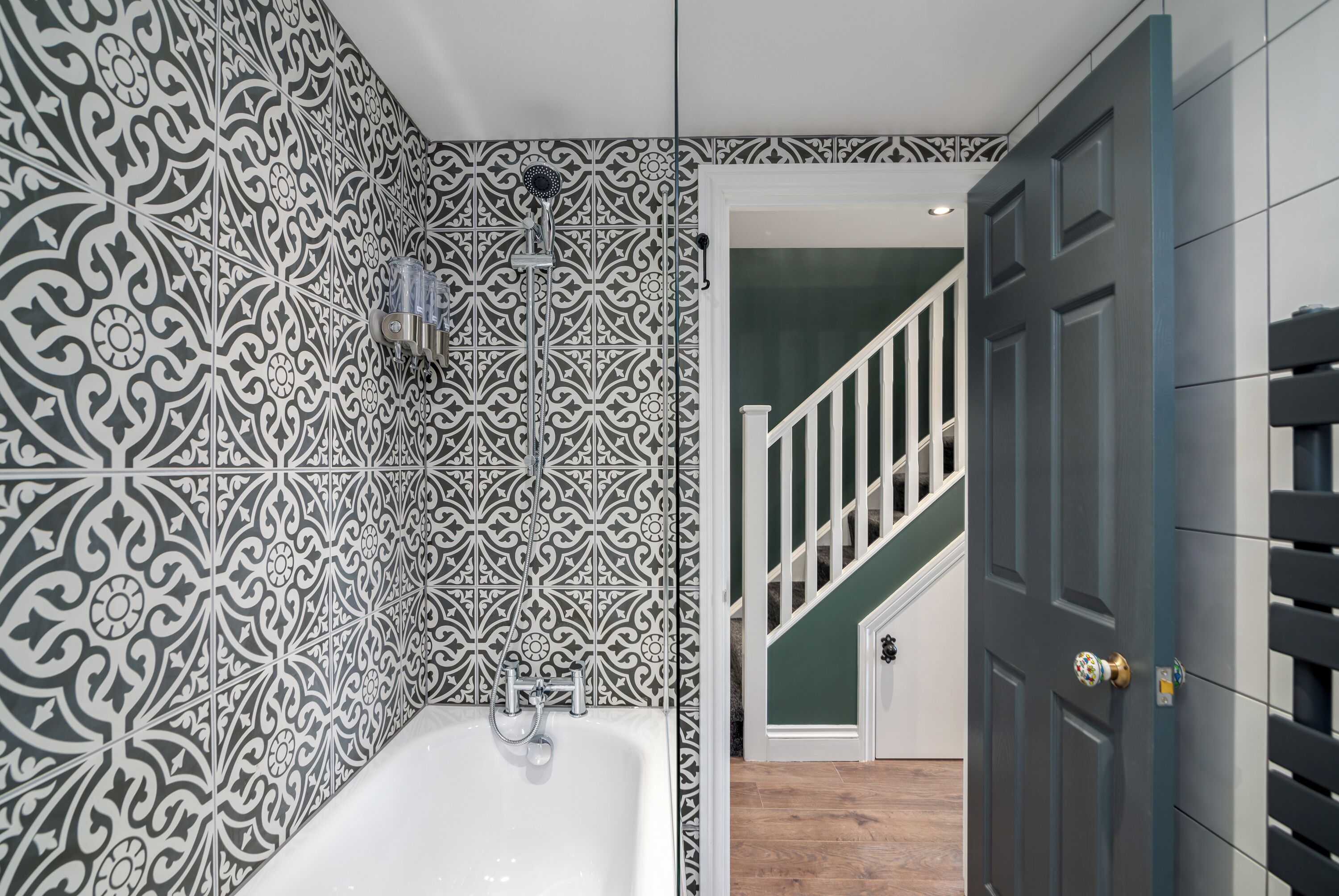 A bathroom with a white bathtub, patterned tiles, a showerhead, and a door leading to a staircase.