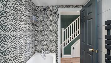 A bathroom with a white bathtub, patterned tiles, a showerhead, and a door leading to a staircase.