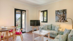 Ein Wohnzimmer mit einer Couch, einem Fernseher, einem Tisch und Stühlen. Es gibt Fenster mit Vorhängen und ein Bild an der Wand.