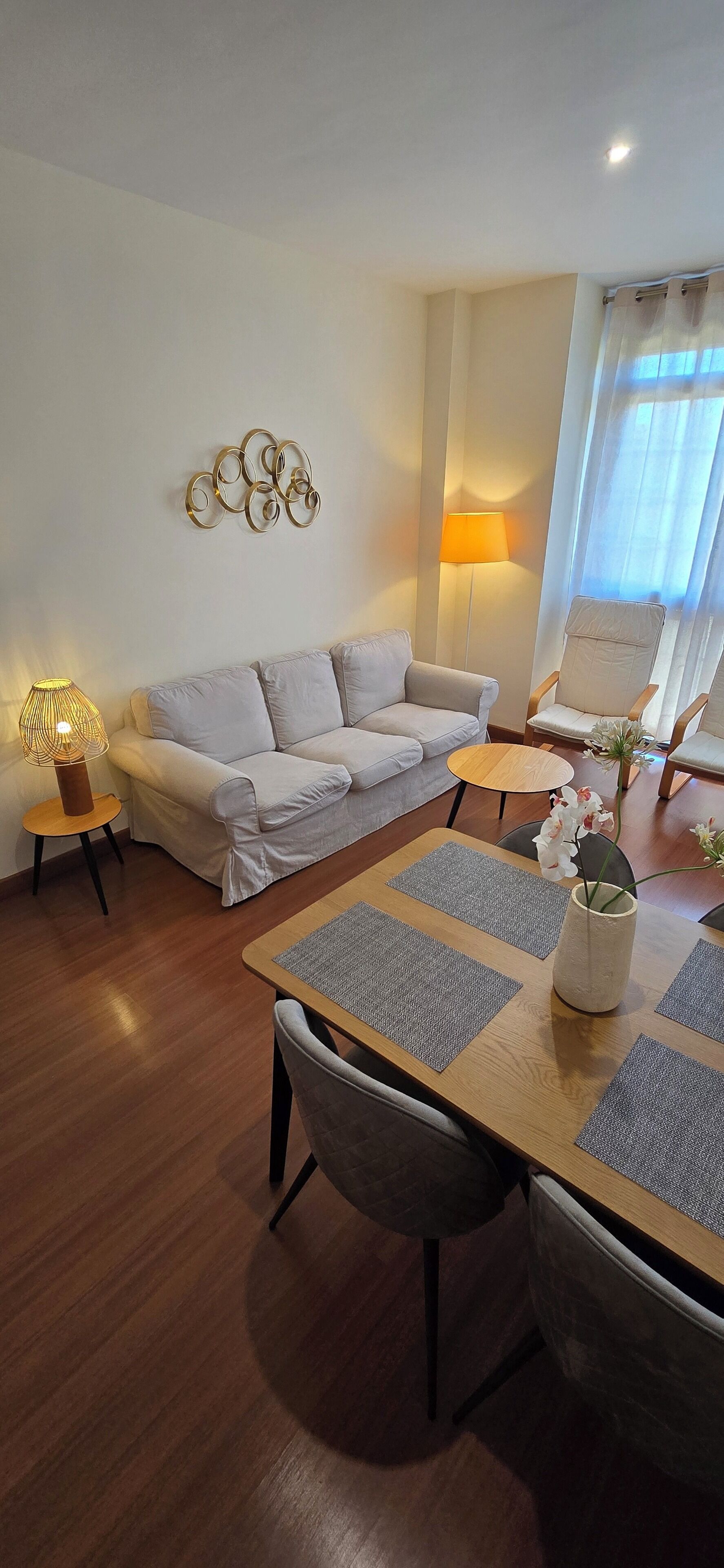 Ein Wohnzimmer mit einer Couch, einem Couchtisch und einem Esstisch mit Platzsets. Eine Lampe steht auf einem Beistelltisch, darüber an der Wand ein dekoratives Wandbild.