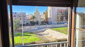 A room with a large window overlooking a street with cars and buildings, including a church. - Ola Rosaleda (Málaga)