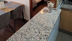 A modern kitchen with granite countertops, a dining area with a table and chairs, and a bedroom visible through an open doorway. - Ola Rosaleda (Málaga)
