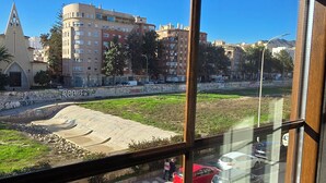 A view through a window of a parking lot with multiple cars, a grassy area, and buildings in the background. - Ola Rosaleda (Málaga)