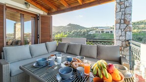 En balkon med en fletsofa, spisebord og morgenmadsbord med frugt, kager og drikkevarer.
