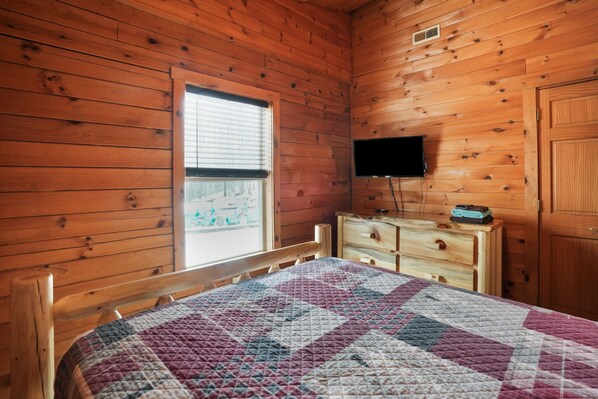 Una habitación de cabaña de madera con una cama, un tocador, un televisor y una ventana con persianas.