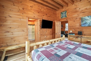 Una habitación tipo cabaña de madera con una cama, un televisor montado en la pared y un banco de madera.