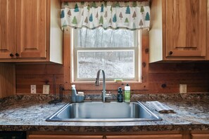 A kitchen with a granite countertop, stainless steel sink, wooden cabinets, and a window with a forest view. - Ridgeview Retreat | Hocking Hills, Hot Tub, Sauna (South Bloomingville)