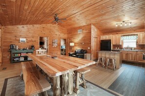 Interior de una cabaña de madera con una gran mesa de comedor, una zona de cocina y un espacio de estar con chimenea.