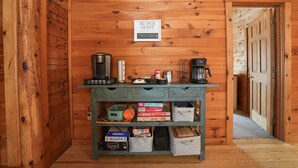 A wooden interior with a sign reading "BE OUR GUEST" and a phone number. A table with board games and storage bins is in the center.