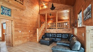 A wooden cabin interior with a leather sectional sofa, a staircase, and a large window.