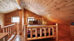 A wooden cabin bedroom with a bed, a window, a dresser, and a wooden railing.
