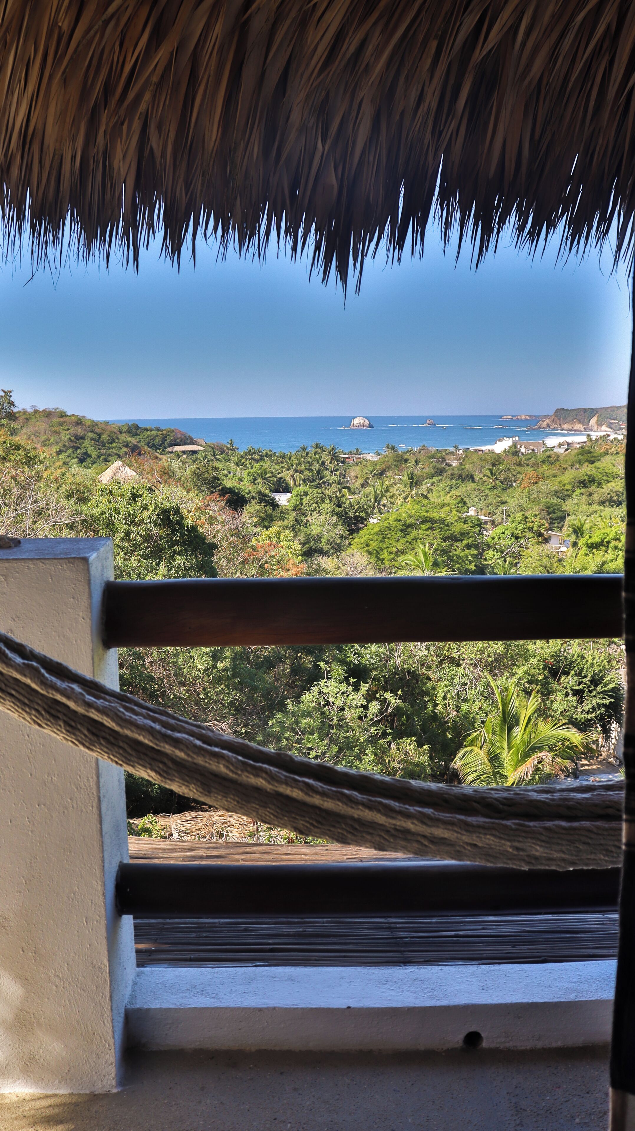 A view from a balcony with a thatched roof, overlooking a coastal landscape with greenery and rocky outcrops.