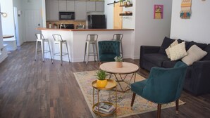 Un salon moderne avec un canapé foncé, des fauteuils turquoise, une table basse et une cuisine en arrière-plan.
