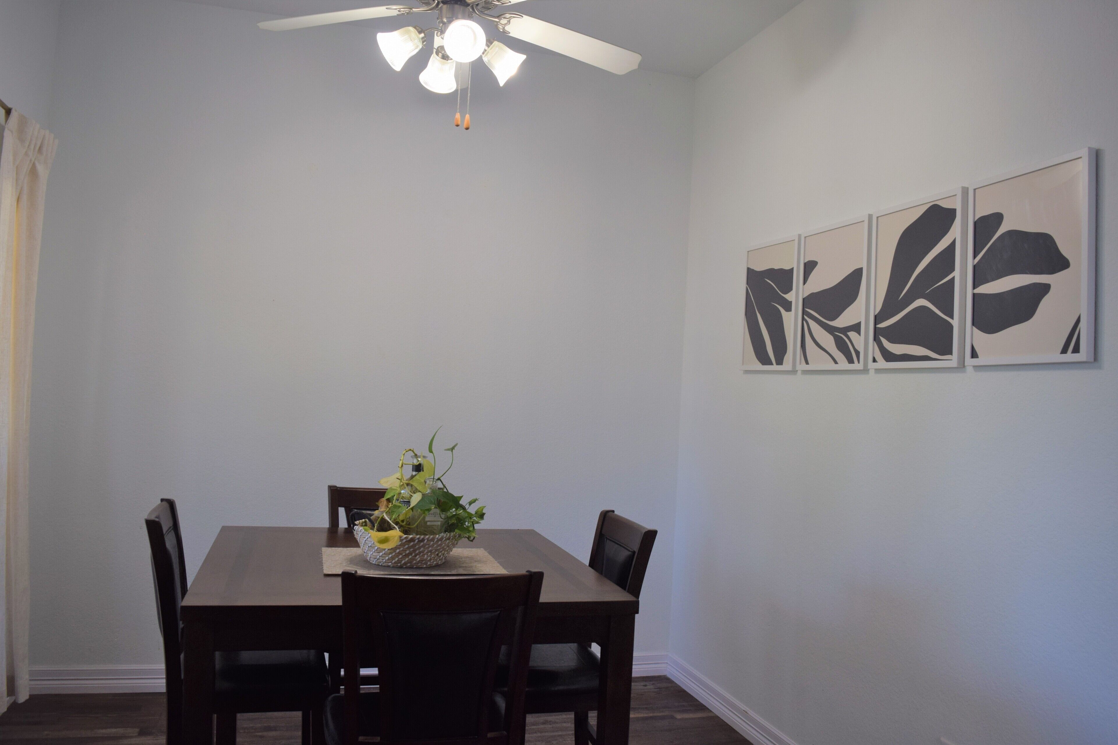 A dining room with a wooden table, four chairs, a ceiling fan, and three framed artworks on the wall.