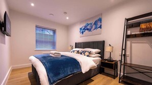 Un dormitorio moderno con una cama grande, una mesita de noche con lámpara, un televisor montado en la pared y una ventana con persianas.