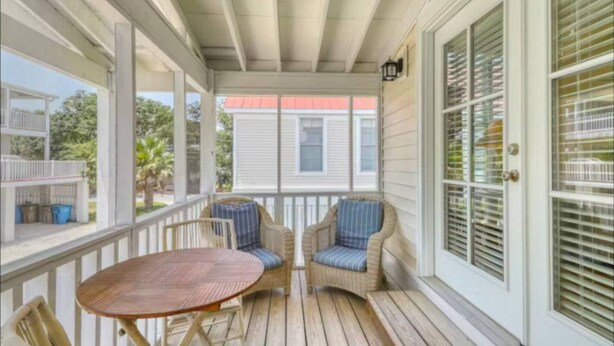 A covered porch with a wooden table and two wicker chairs, a white door with blinds, and a view of a red-roofed building. - Bay Breeze - 3 Min Walk to Beach (Tybee Island)