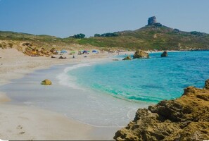 Ein Strand mit klarem türkisblauem Wasser, einem sandigen Ufer und einer Burg auf einem Hügel im Hintergrund.