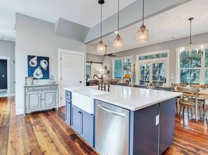 A modern kitchen with a central island, wooden flooring, pendant lights, and a dining area with wooden chairs. - Marsh front/sleeps 8/ Walkable/ Rec Area (Tybee Island)