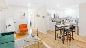 A modern kitchen with white cabinets, a dining area with a glass table, and a bright, open space with a green sofa and an orange chair. - City Center Oasis Newark (Newark)