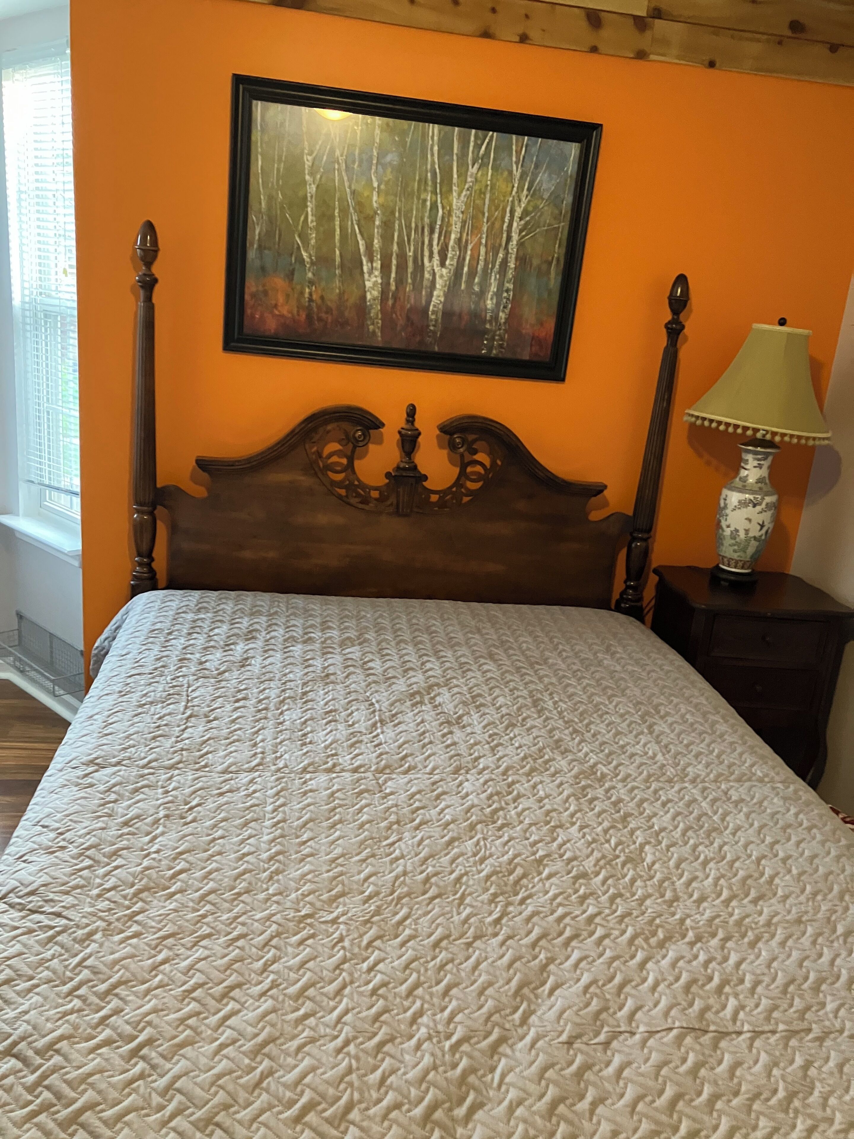 A bed with a textured white cover, a wooden headboard, and a painting of trees on the wall.