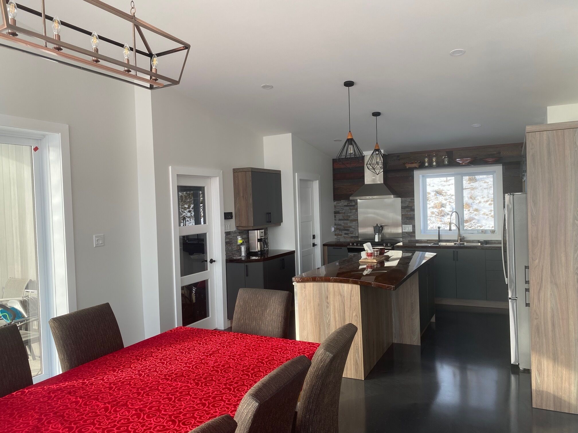 Una cocina moderna con una isla central, un comedor con mantel estampado rojo y vistas a un paisaje nevado a través de una ventana.