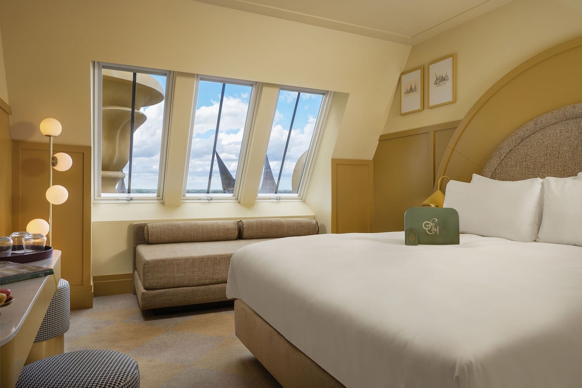 A hotel room with a large bed, a seating area, a desk with a lamp, and a window with a view of the sky.