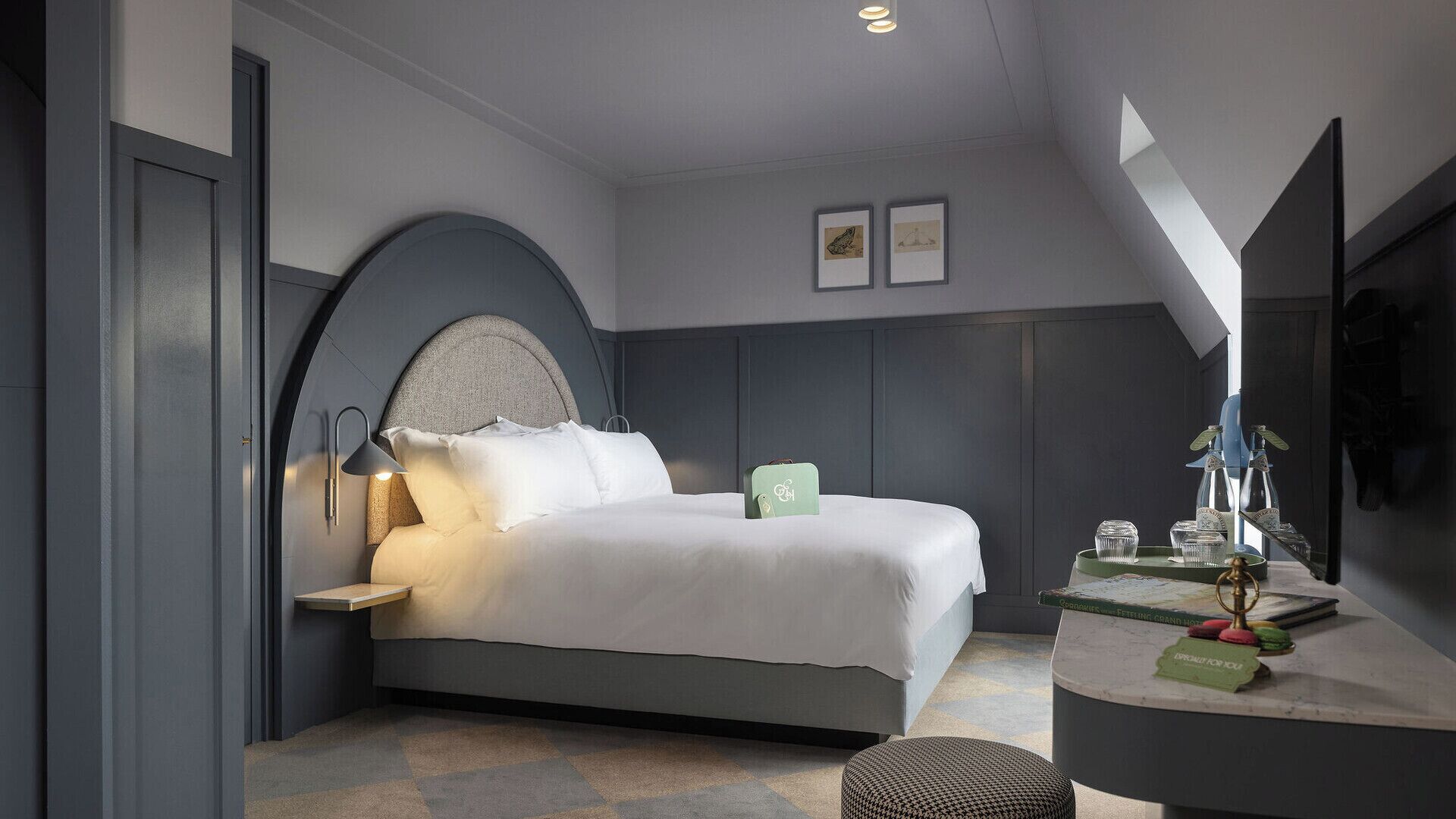 Une chambre d’hôtel moderne équipée d’un lit, d’une table de chevet, d’une lampe et d’un petit bureau sur lequel se trouve un plateau avec divers objets.