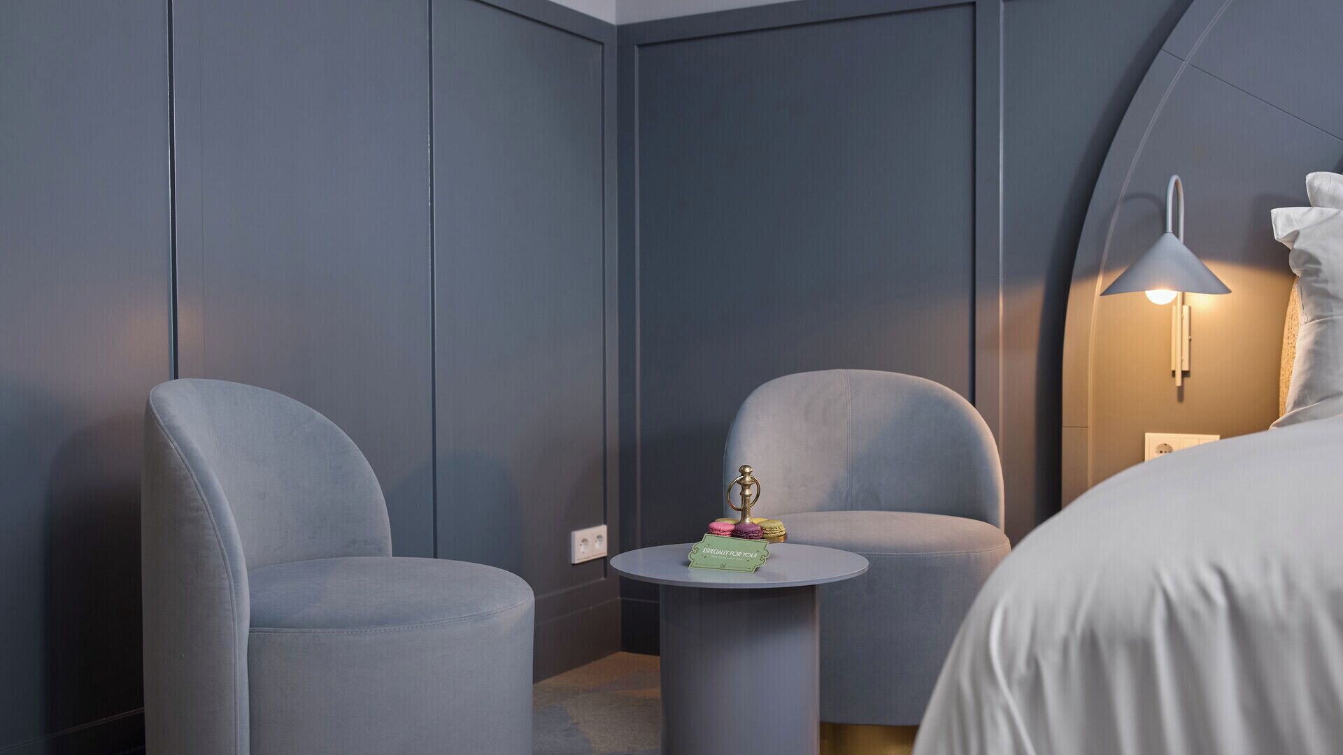 Une chambre d’hôtel moderne avec deux fauteuils gris, une petite table ronde agrémentée d’un objet décoratif et un lit recouvert d’une couverture blanche.